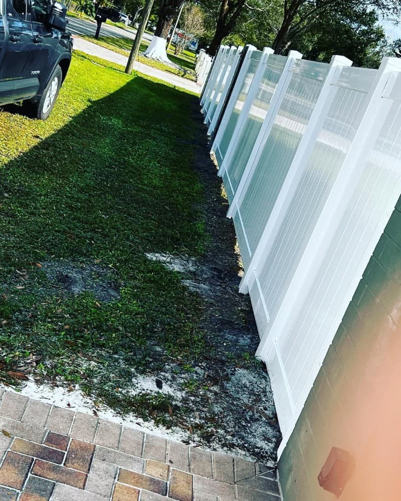 Diagonal white vinyl privacy fence long run pavers corner grass mulch truck distant trees suburb bright sunlight shadow.
