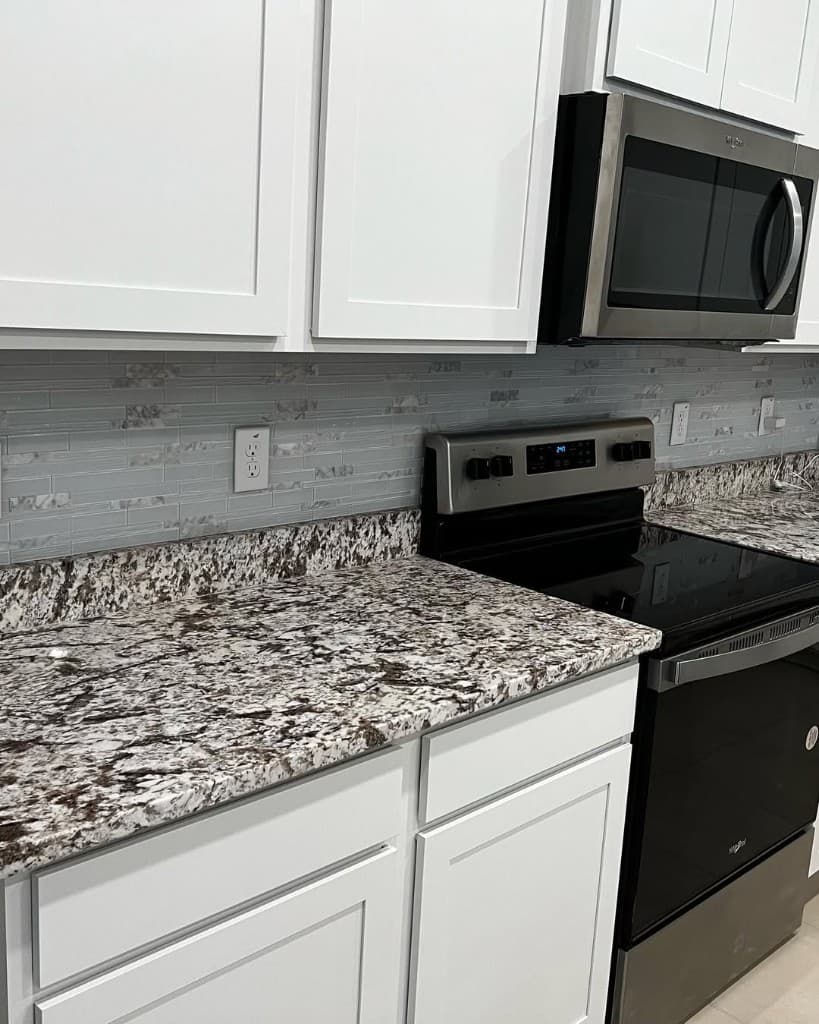 Angled kitchen view with white cabinets, glass linear backsplash, granite counter, stainless stove, and microwave.