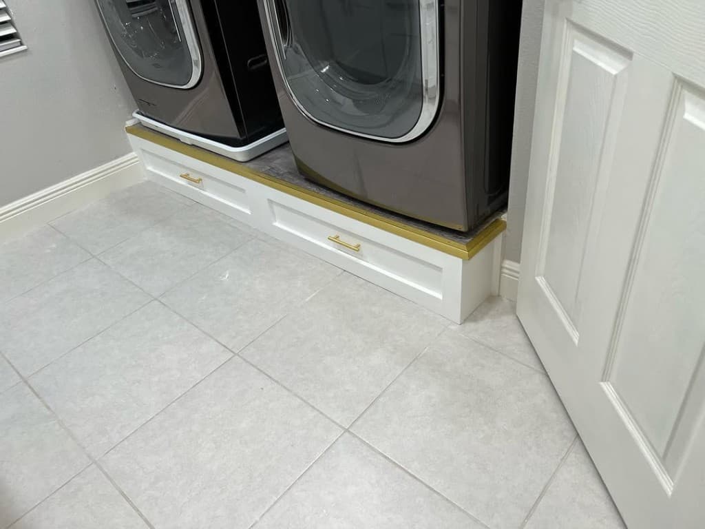 Stacked washer dryer white painted pedestal shaker drawers brass handles gold trim ornate header grey tile floor doorway.