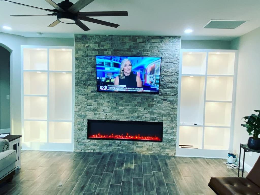 Great room ledger stone accent TV electric flame white built shelving cubes puck lights ceiling fan plank floor sofa.