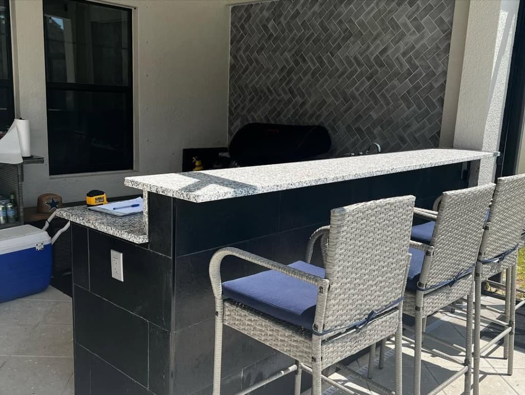 Covered patio outdoor kitchen with two-level granite bar, herringbone tile wall, wicker bar stools, and stainless sink area.