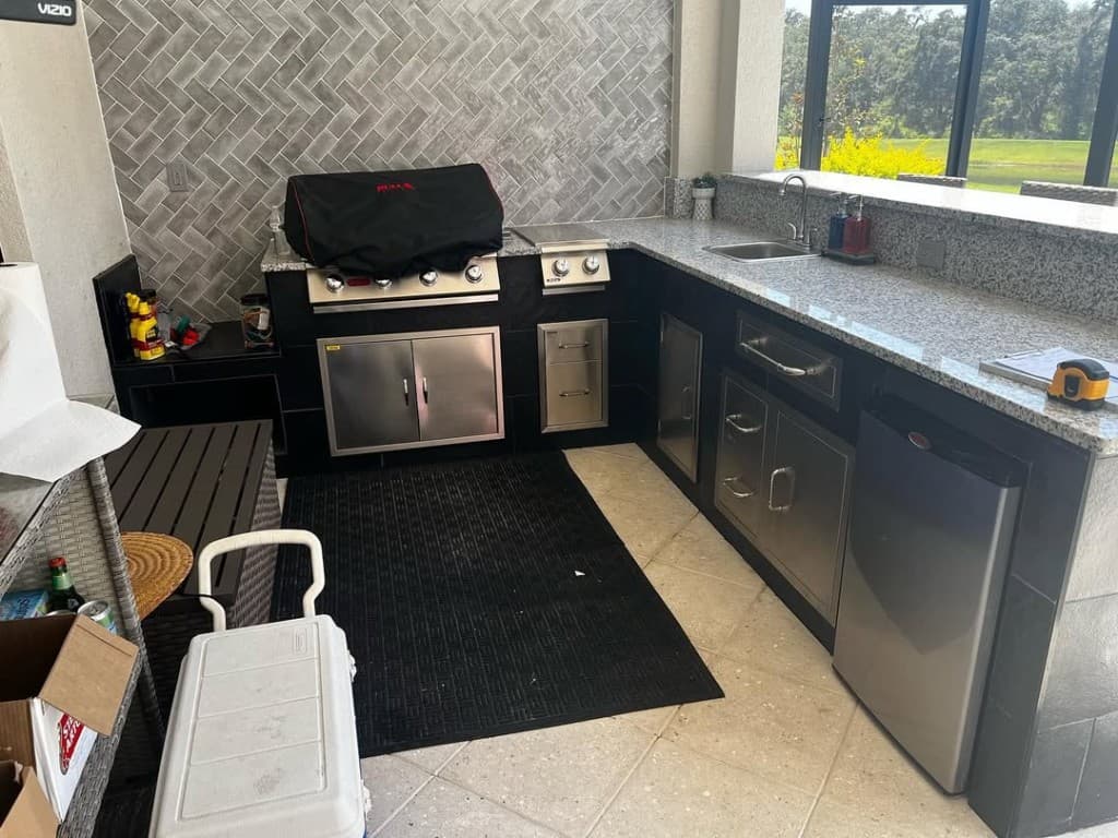 L-shaped outdoor kitchen with black cabinetry, grey herringbone tile, granite counters, BULL grill cover, and patio tile floor.