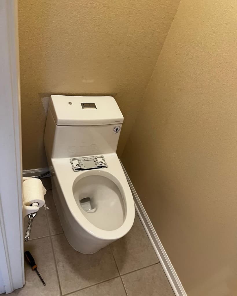 White one-piece toilet without seat wrench screwdriver beige tile dual flush buttons wall patch behind tank bathroom.